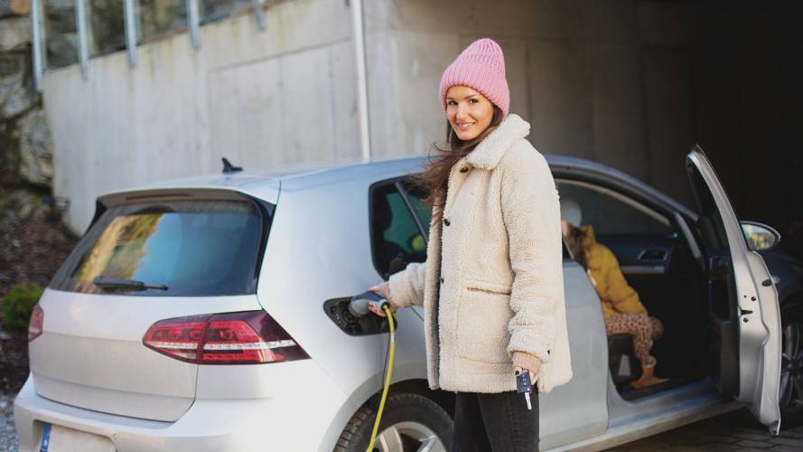 Smilende dame i lue og vinterjakke som holder i ladekabel som er koblet i elbil