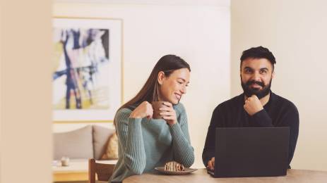 Ungt smilende par sitter ved kjøkkenbordet med kaffekopp og laptop