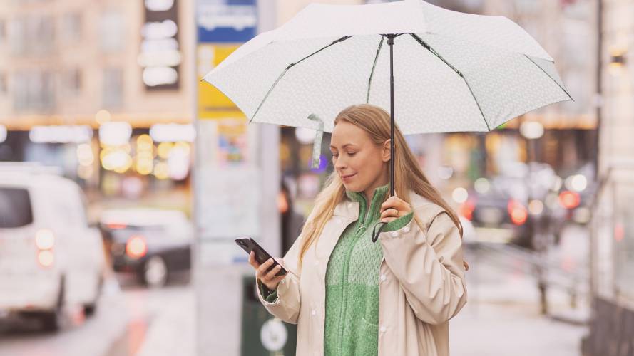 Ung kvinne med mobil og paraply venter på buss