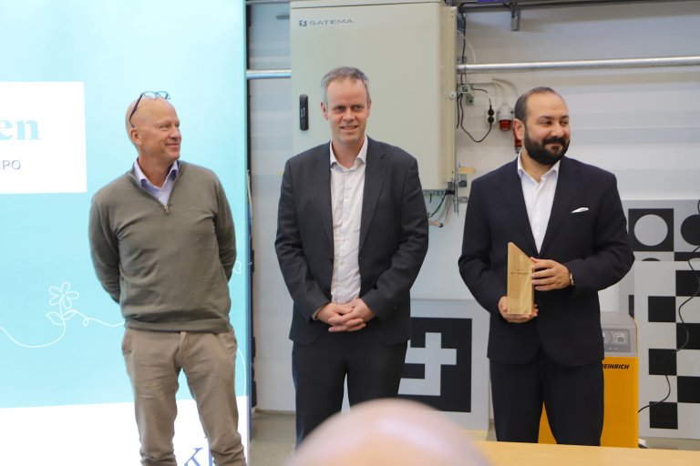 Tre menn smiler foran forsamling.