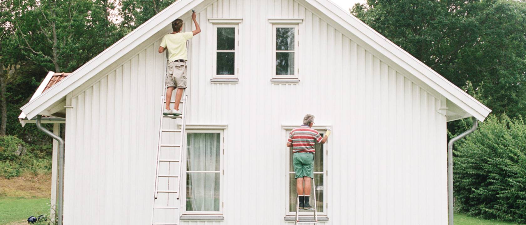 To menn som står på  hver sin stige og maler fasaden på et hvitt trehus i shorts og t-skjorte