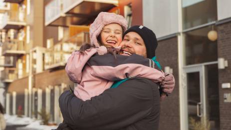 Smilende far og datter i vinterklær som klemmer hverandre med boligblokker i bakgrunnen