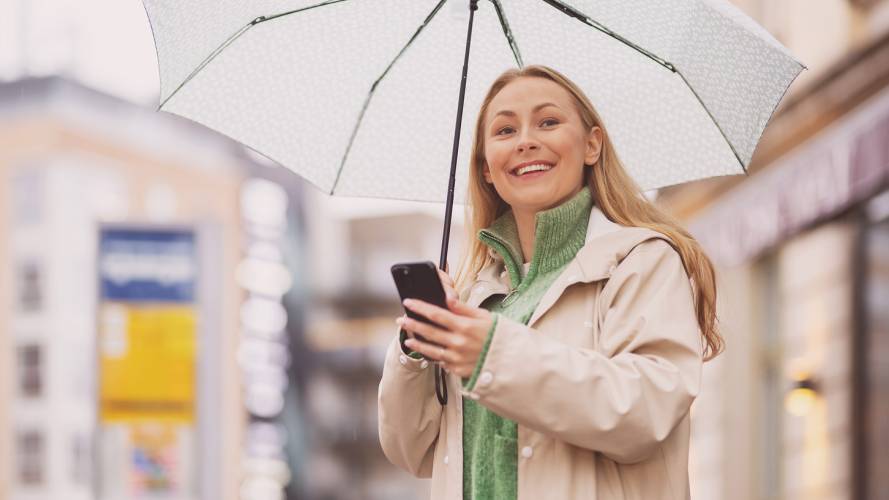 Smilende ung kvinne med mobil og paraply venter på buss