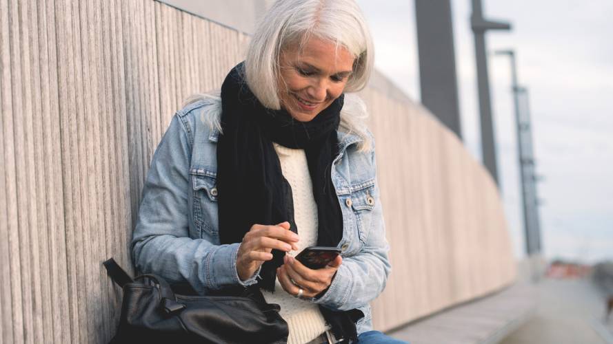 Smilende dame i jeansjakke og skjerf som sitter på benk ute med mobilen i hånden og veske ved siden av seg