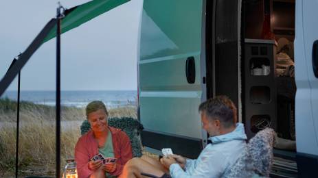 Smilende dame og mann som spiller kort utenfor campingbil på en sommerkveld