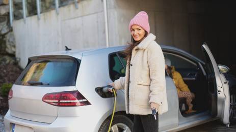 Smilende dame i lue og vinterjakke som holder i ladekabel som er koblet i elbil