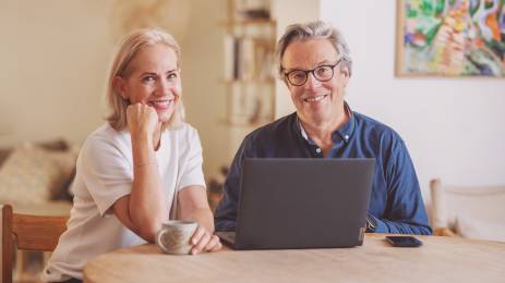 Voksen kvinne og mann med laptop sitter ved kjøkkenbordet og smiler